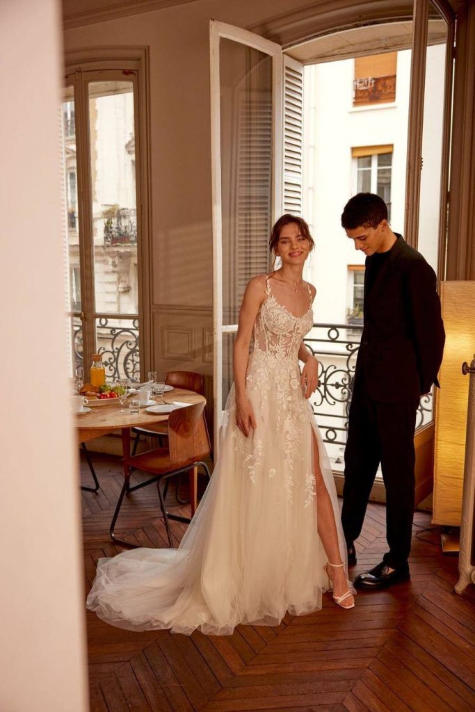 Bride in a floral applique gown with a high slit standing with her partner in a sunlit apartment