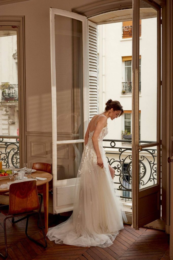 Bride in a floral lace A-line gown standing at open balcony doors in warm afternoon light