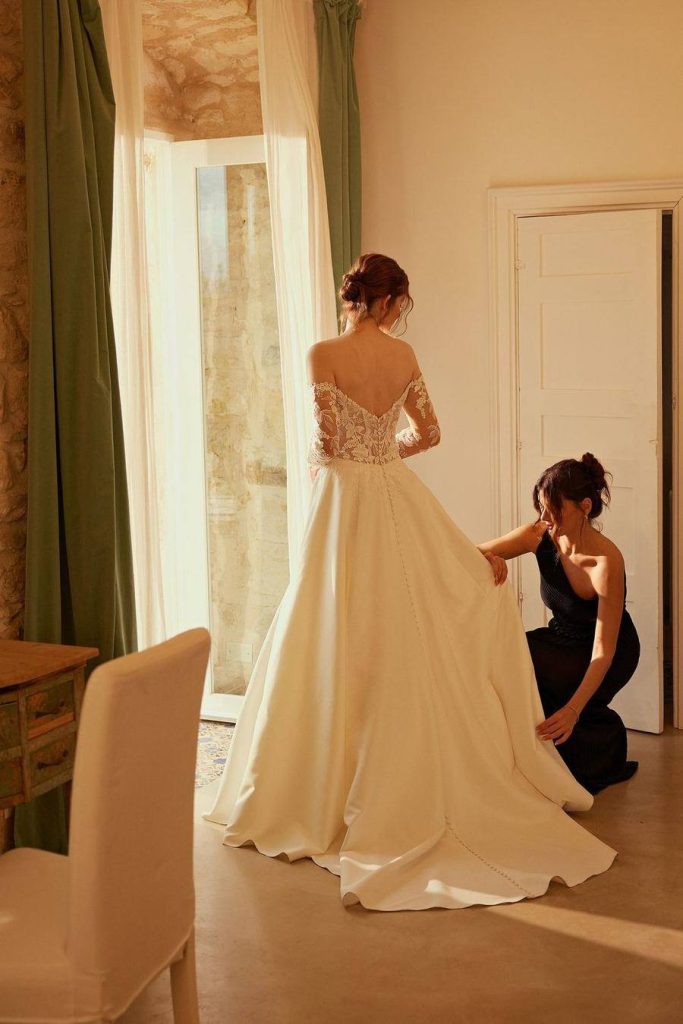 Stylist adjusting the train of an off-the-shoulder lace and satin gown beside a tall window